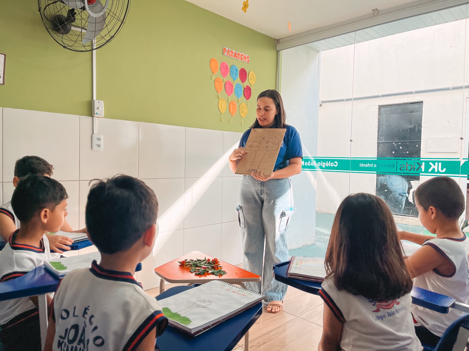 Professora apresenta o desenho de uma árvore à turma, com folhas sobre a mesa